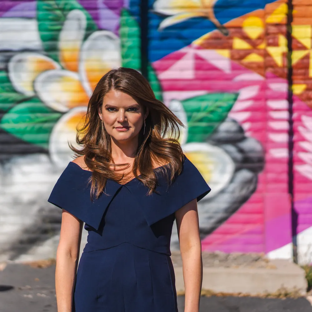 Lindsey Elmore in a navy dress in front of a brightly painted mural, looking straight at the camera