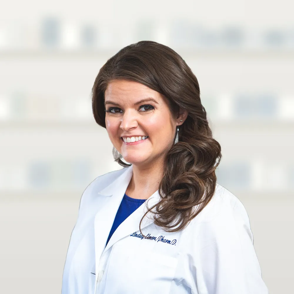 Lindsey Elmore smiling in a white pharmacist's coat with her name embroidered on it, in front of softly blurred white pharmacy shelves